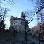 Bran Castle, Romania Ghost Tours- castle on hill, trees, blue sky, flag down castle wall, Romania