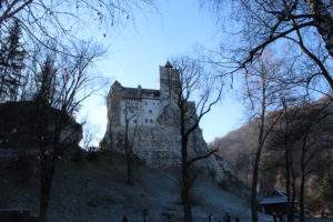 Bran Castle, Romania Ghost Tours- castle on hill, trees, blue sky, flag down castle wall, Romania