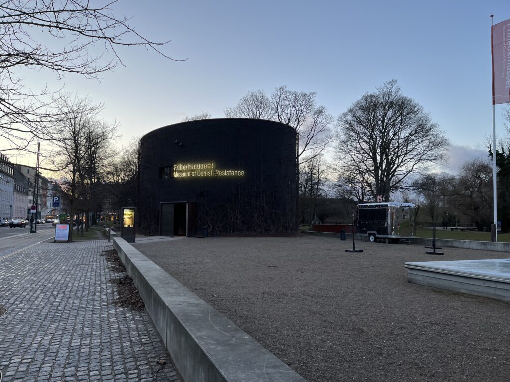 Under the cloak of darkness, the Resistance Museum in Copenhagen stands as a silent guardian, illuminating the stories of bravery and defiance that echo through the night. At the side of the carpark you will see the circular museum and a silver hot dog truck