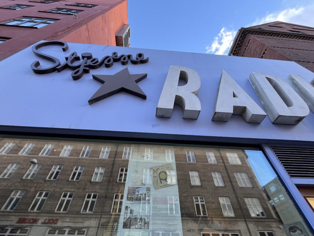 The blue sign with black and white writting and a black star stands above the window full of old photographs of the Danish resistance members who used to use this radio shop as a location
