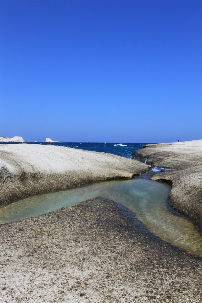 blue skies overlook the blue waters and sandy and pebbled beach on the Island of Milos